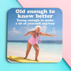 Funny woman's coaster; featuring an old lady, standing on a surfboard in the summer sun; bold white text behind her reads 'old enought o know better, young enough to make a tit of yourself anyway'; by Dean Morris.