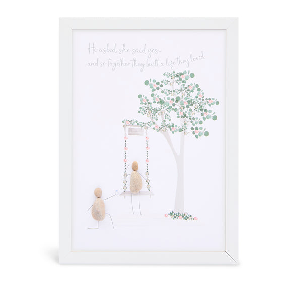 This white shadowbox framed pebble art A4 print features a person swinging on a swing under a tree and another on bent knew proposing.&nbsp; It reads He asked she said yes... and so together they built a life the loved.

This would be a great gift for an engagement, wedding, or an anniversary.