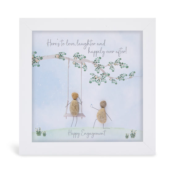 This white shadowbox framed pebble art print features someone on swing hanging from a tree with someone else pusing them.&nbsp;It reads "Here's to love, laughter and happy ever after! Happy Engagement!