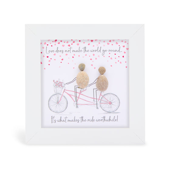 This white shadowbox framed pebble art print features a lovely couple on romantic tandom bike ride together with flowers in the basket on the front of the bike and read hearts floating above.&nbsp;It reads Love does not make the world go 'round, it makes the ride worthwhile.