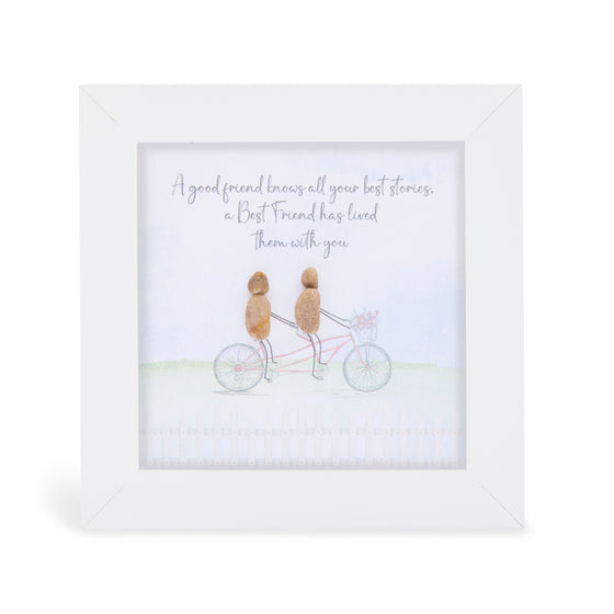 This white box framed pebble art print features two friends riding a bike together with flowers in the basket on the front of the bike. It reads A good friend knows all your best stories, a best friend has lived them with you.